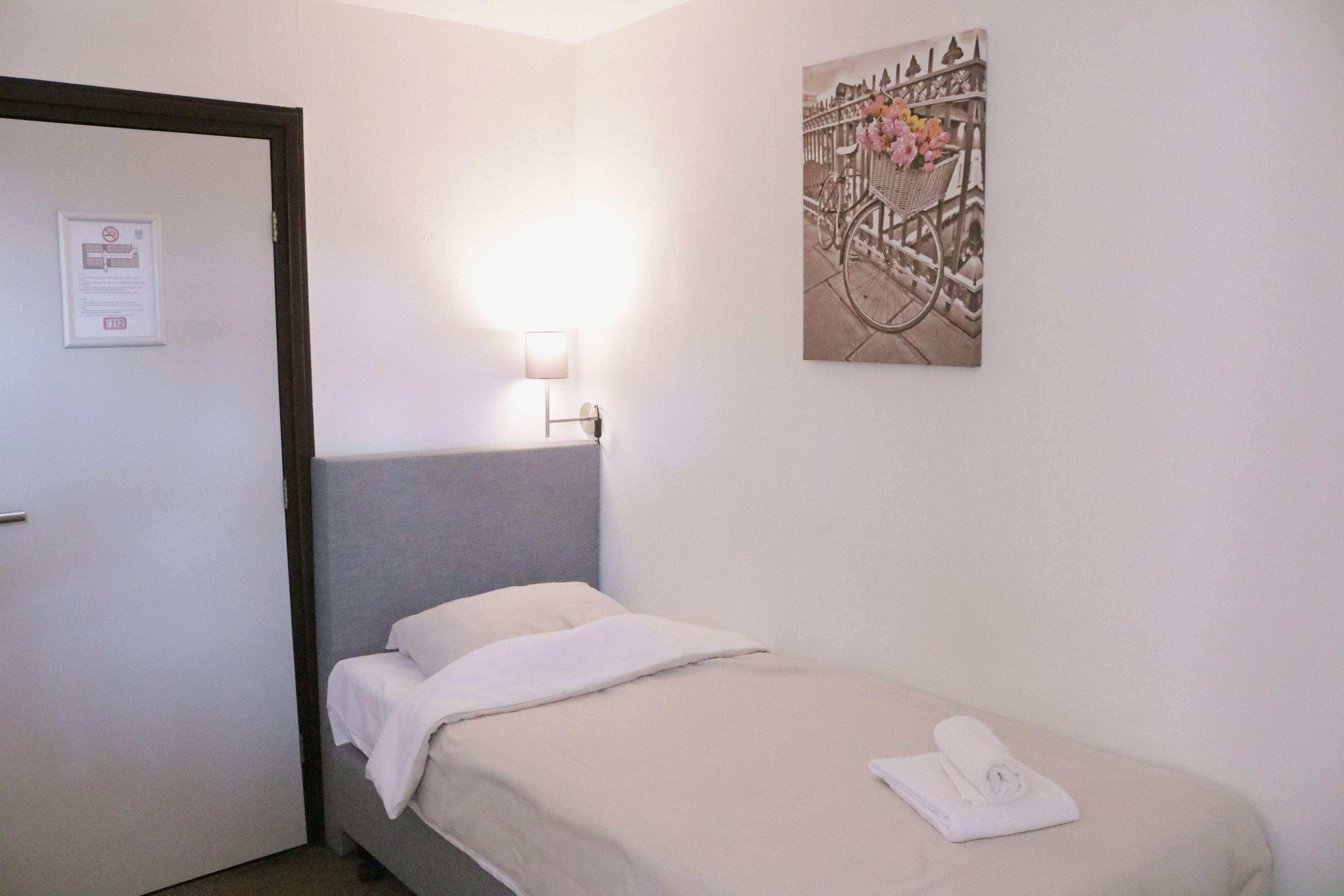Uma cama pequena e bem arrumada com travesseiro branco e uma toalha enrolada, uma luminária fixada na parede e um quadro com uma bicicleta e flores.
