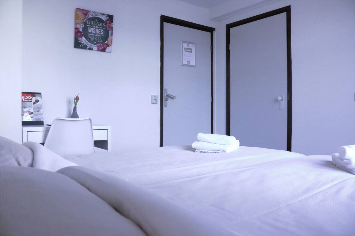 A hotel room with a neatly made bed, a desk with a chair, and two doors.