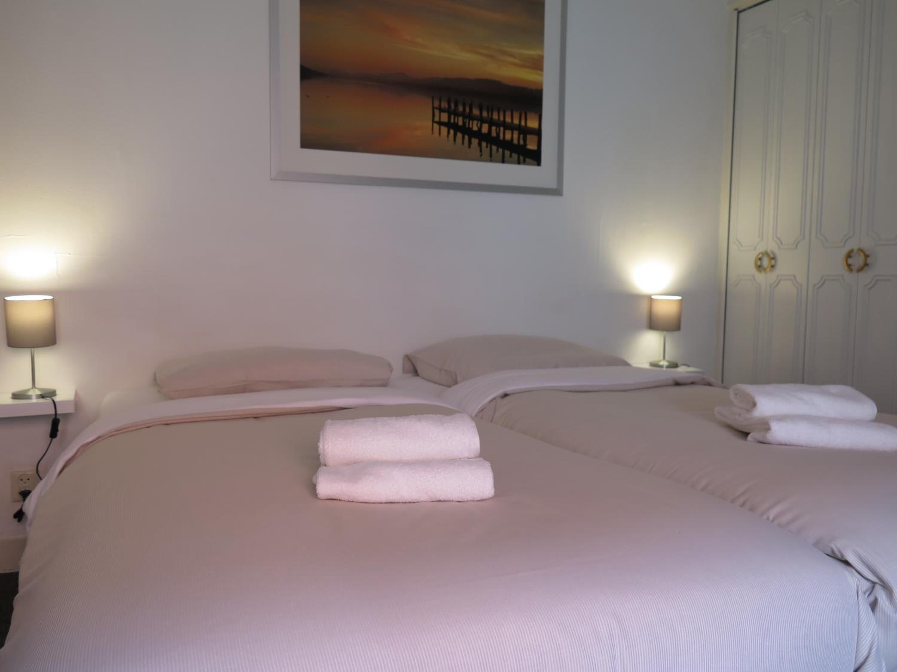 Habitación de hotel con una cama grande, dos lámparas de noche, un cuadro de una puesta de sol y una puerta blanca con pomo dorado.