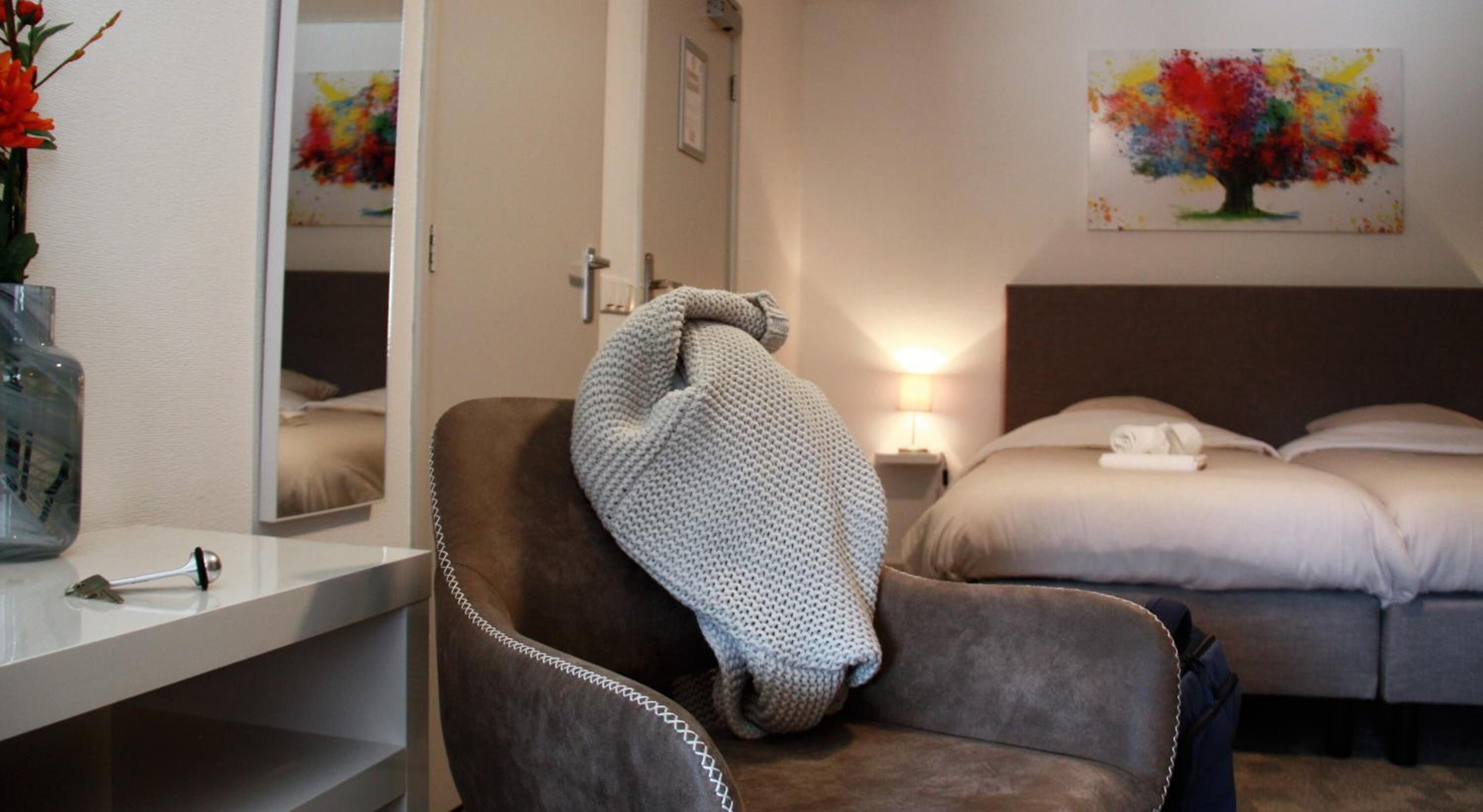 Una camera d'albergo con un letto, una sedia con una coperta, un comodino con una lampada e un quadro appeso al muro.