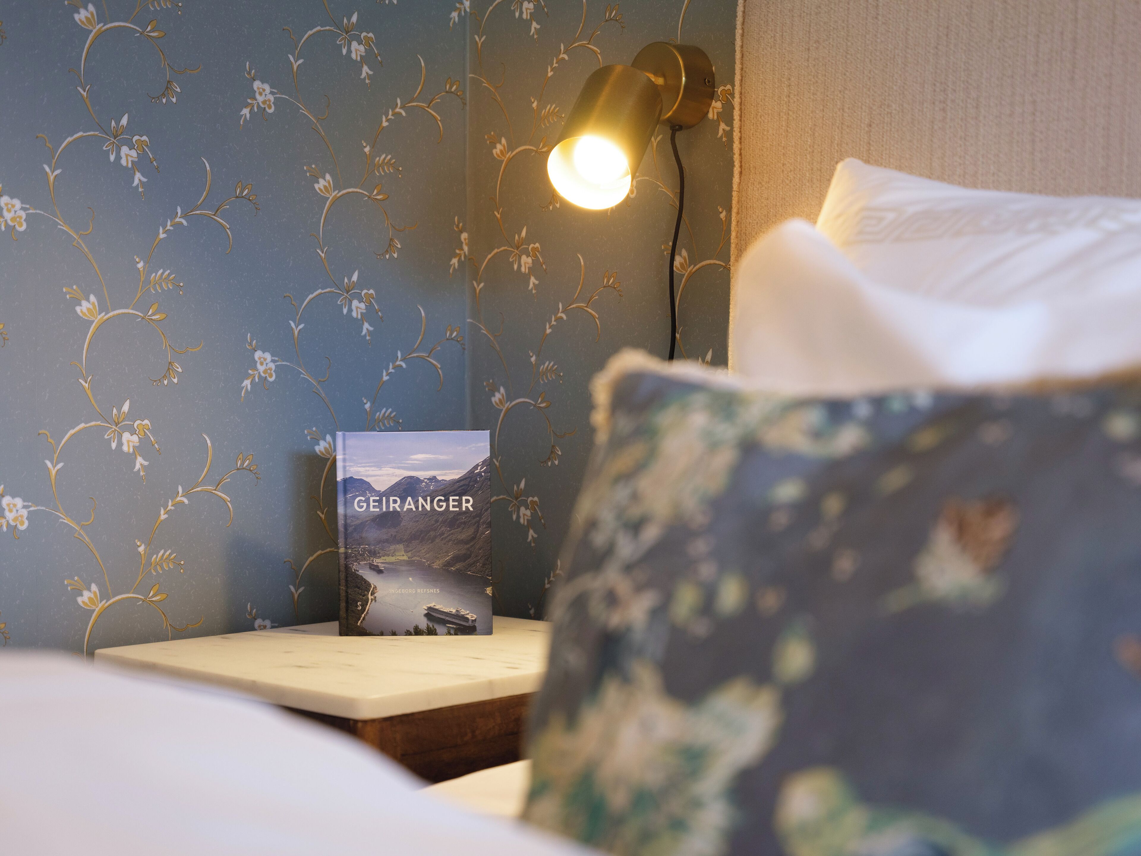 A neatly made bed with a pillow, a bedside table with a book titled 'GEIRANGER', and a wall-mounted lamp.