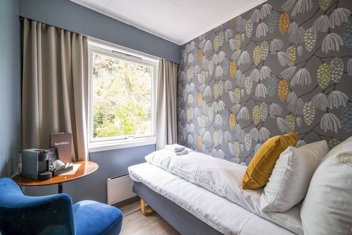A hotel room with a bed, a small table with a coffee maker, a chair, and a window with curtains.