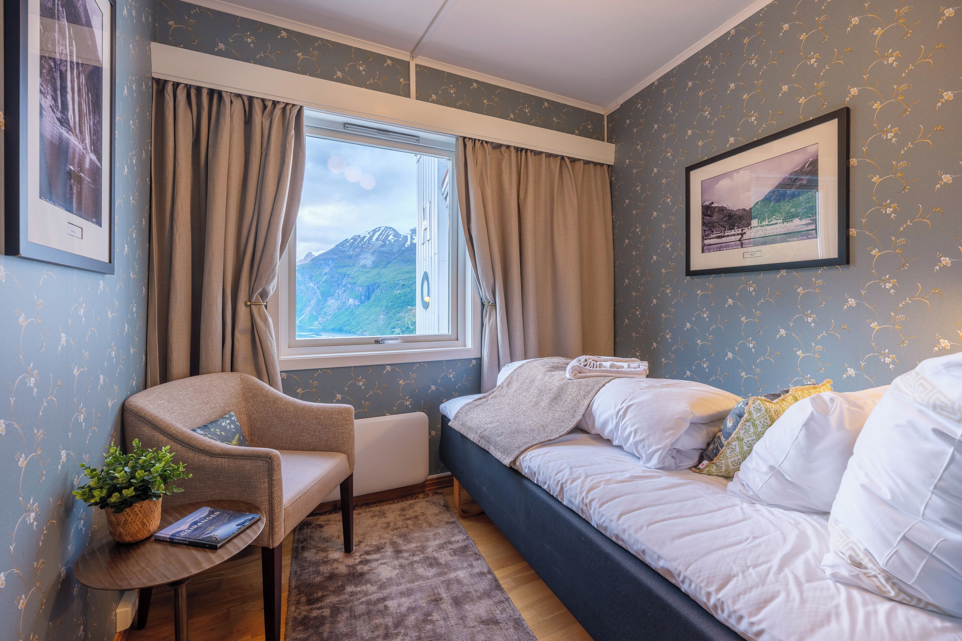 A cozy room with a bed, a chair, a small table with a plant, and a window with curtains.