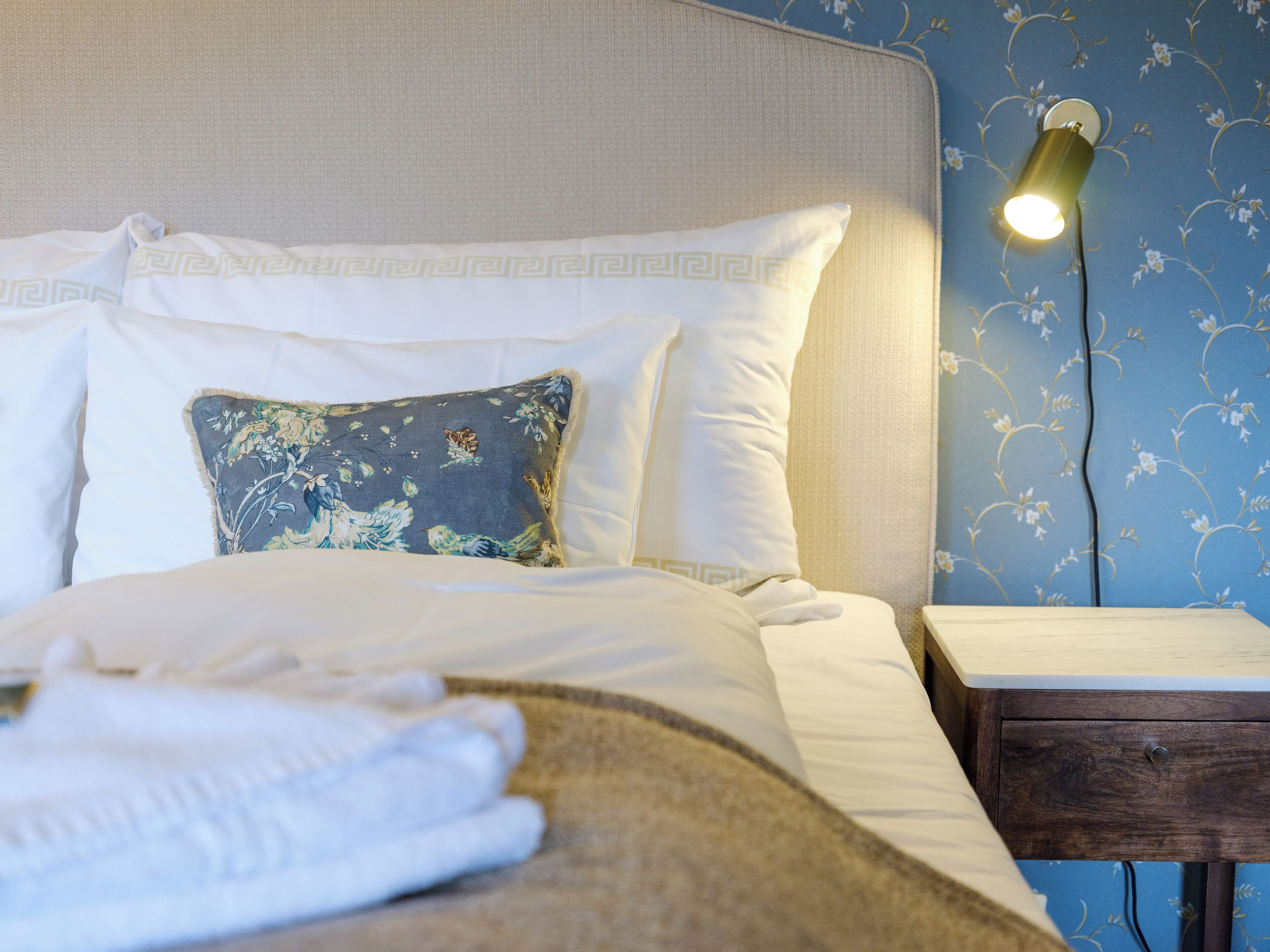 Un lit bien fait, avec du linge de lit blanc et un coussin décoratif, une lampe de chevet et une table de chevet en bois.