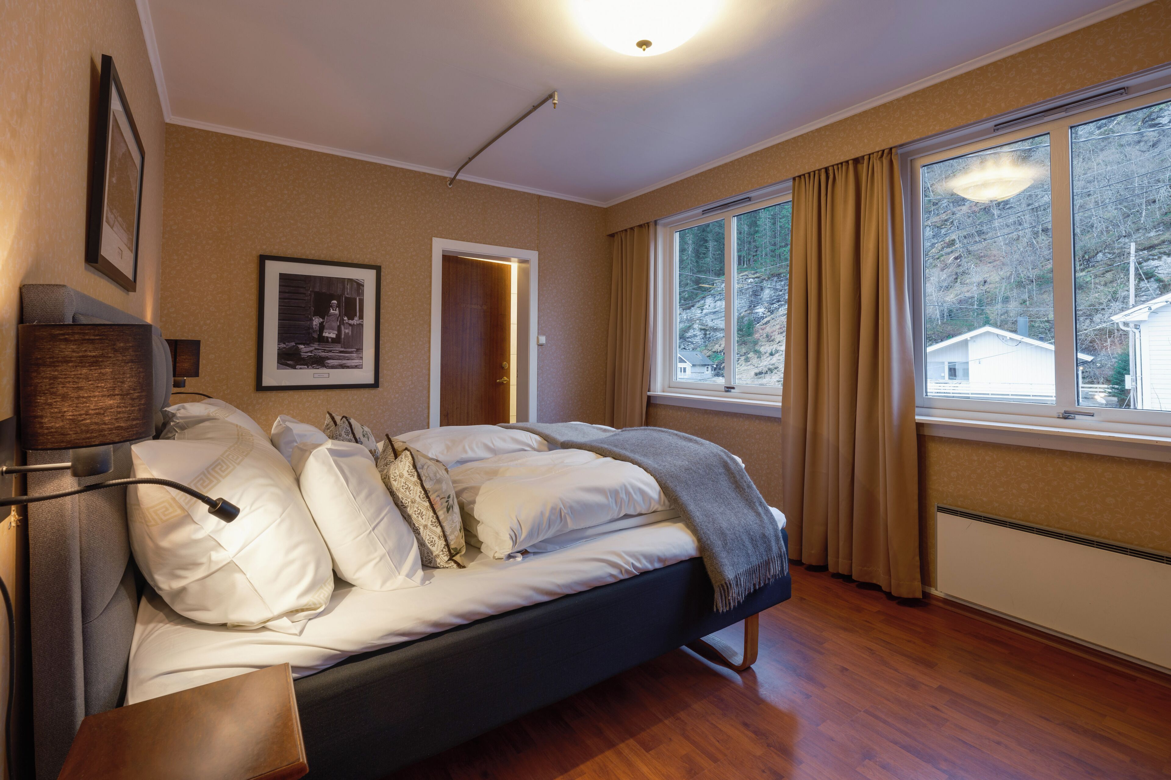 A bedroom with a bed, a bedside table, a lamp, a framed picture on the wall, and a window with curtains.