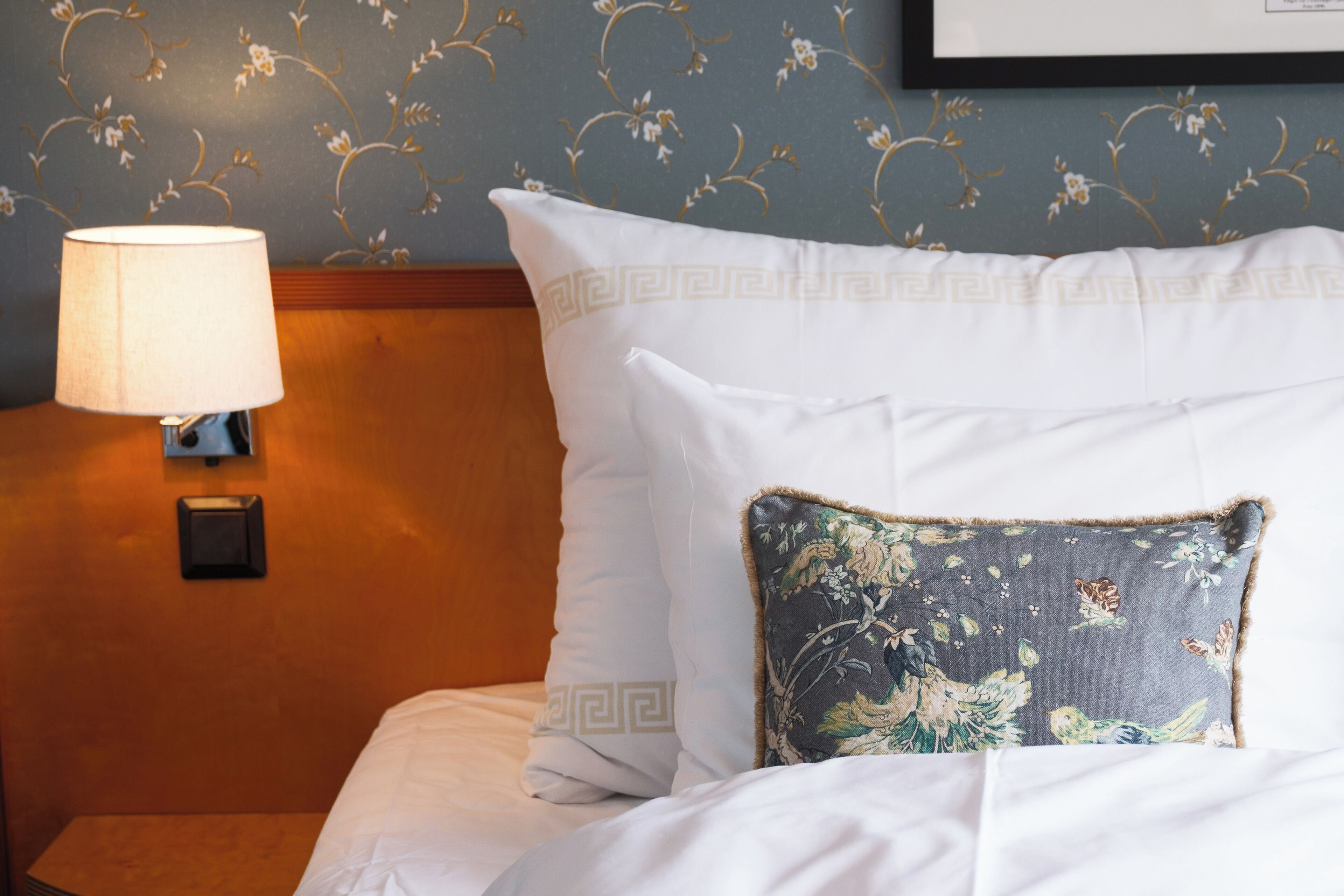 A neatly made bed with white bedding and a decorative pillow, a wall-mounted lamp, and a floral-patterned wall.
