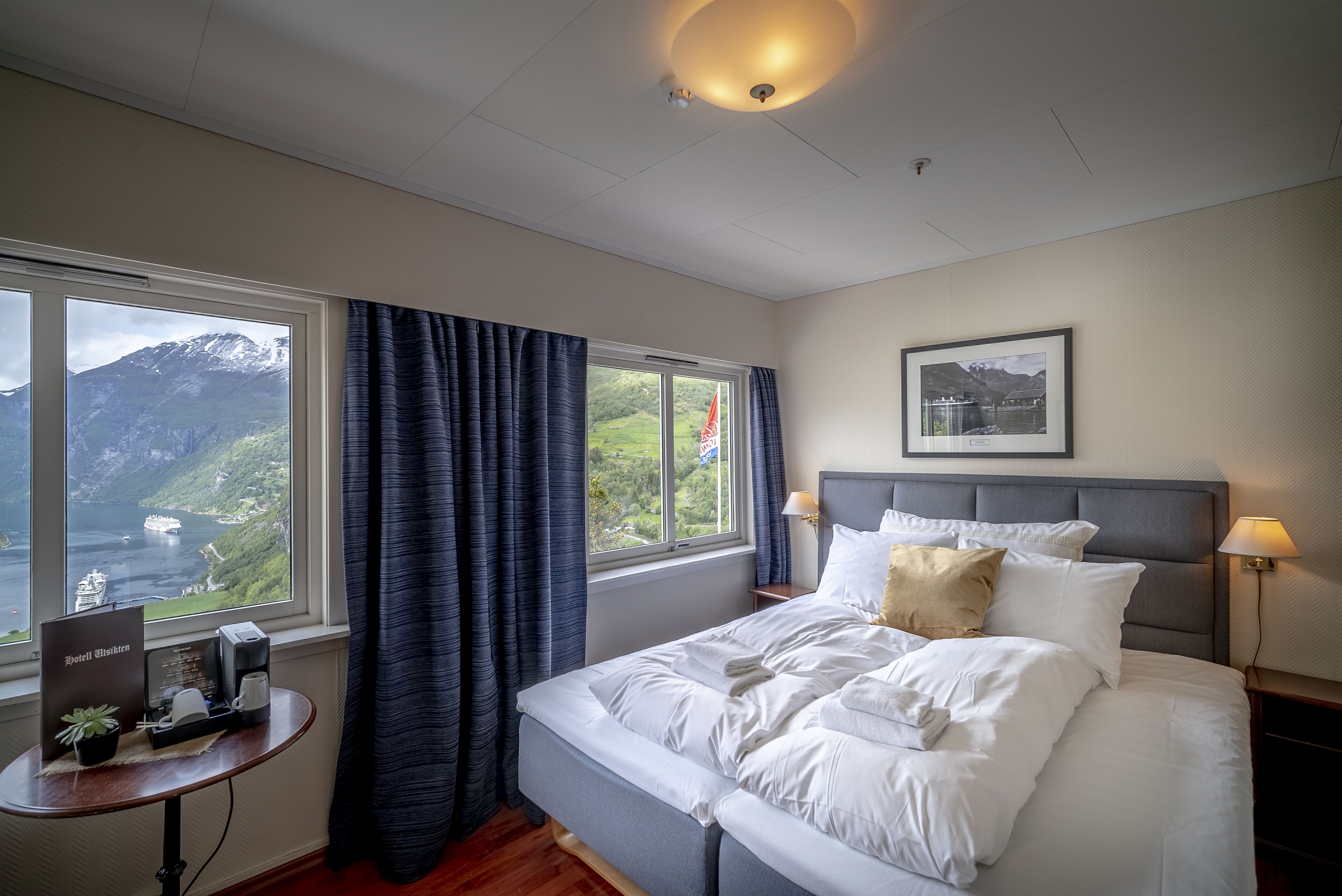 A hotel room with a bed, a bedside table with a coffee maker, a lamp, and a view of mountains and a lake.