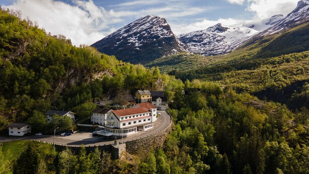 A mountain lodge with a curved road leading to it, surrounded by dense forest and snow-capped peaks.