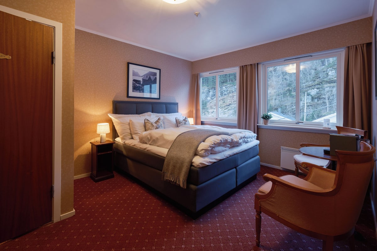 A bedroom with a bed, bedside table, chair, and a window with a view of trees.