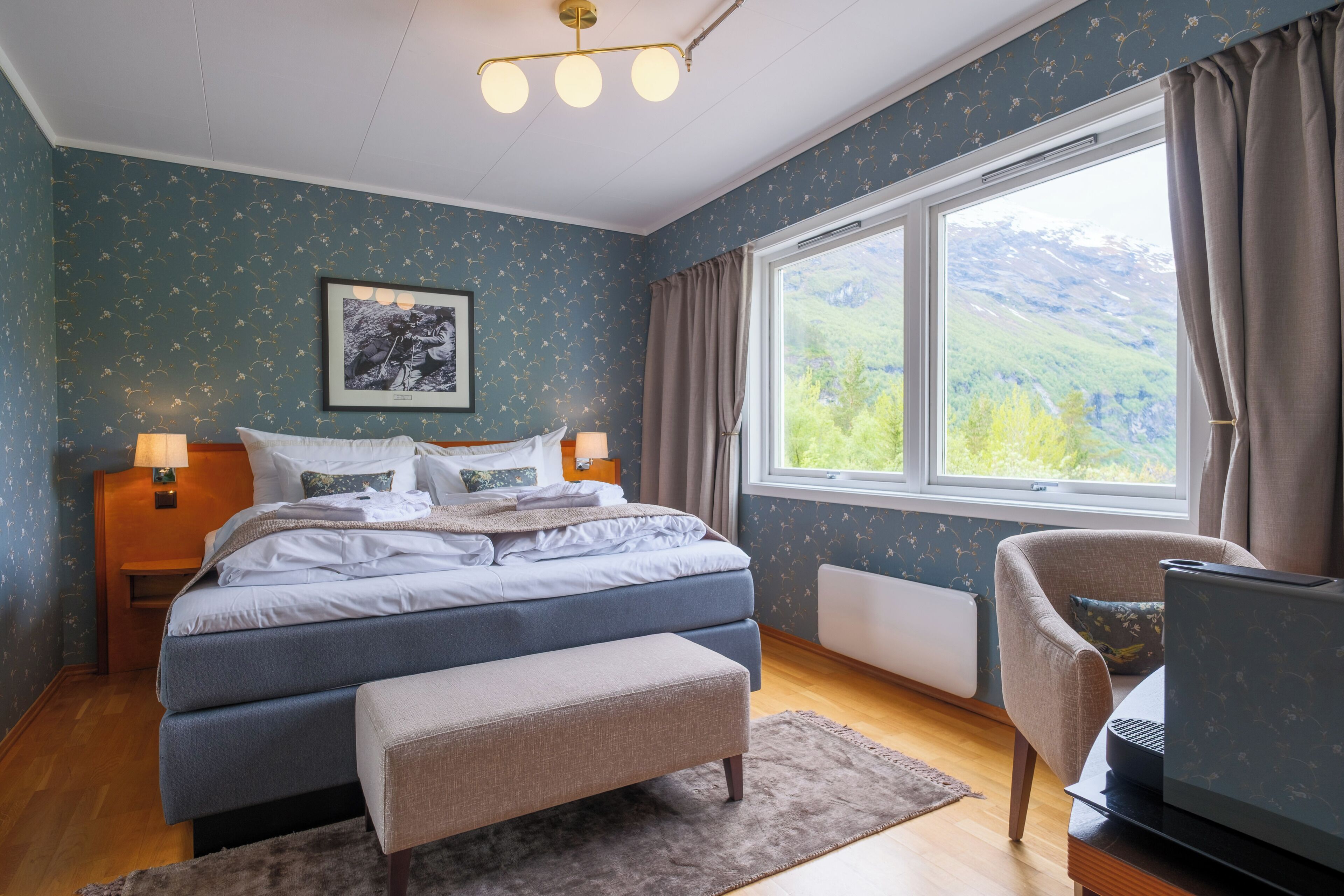 Un dormitorio con cama, mesitas de noche, un escritorio con una computadora portátil, una ventana con vistas a montañas y un cuadro en la pared.