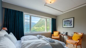 A hotel room with a large bed, two chairs, a small table, a window with a mountain view, and a framed picture on the wall.