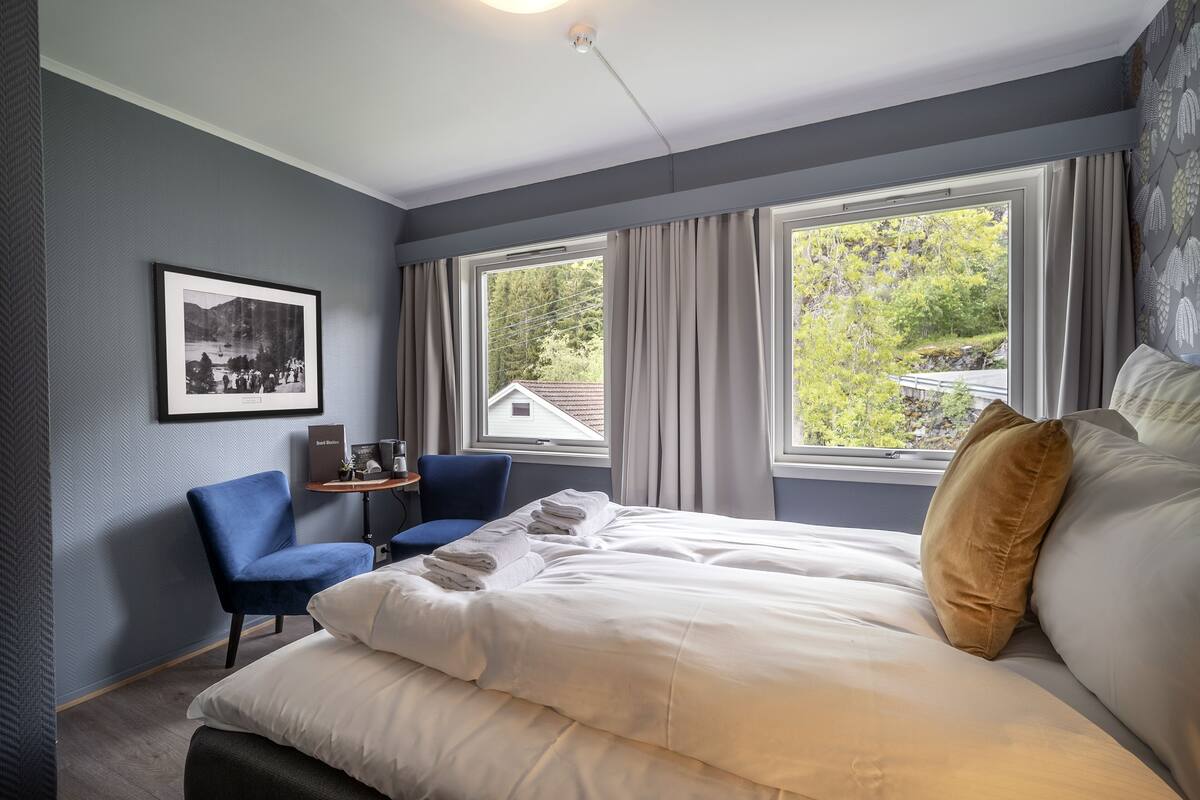 A hotel room with a large bed, a desk with a chair, a framed picture on the wall, and a window with curtains.