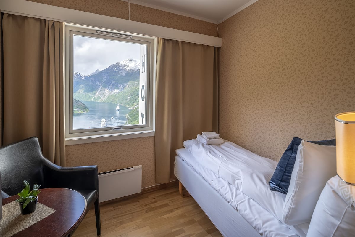 A hotel room with a bed, a table with a plant, a chair, a window with curtains, and a view of mountains and a lake.
