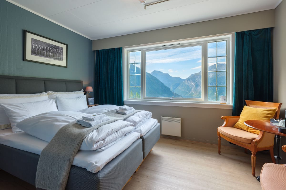 A neatly made bed with white linens and a grey blanket, a wooden chair with a yellow cushion, and a window with a mountain view.