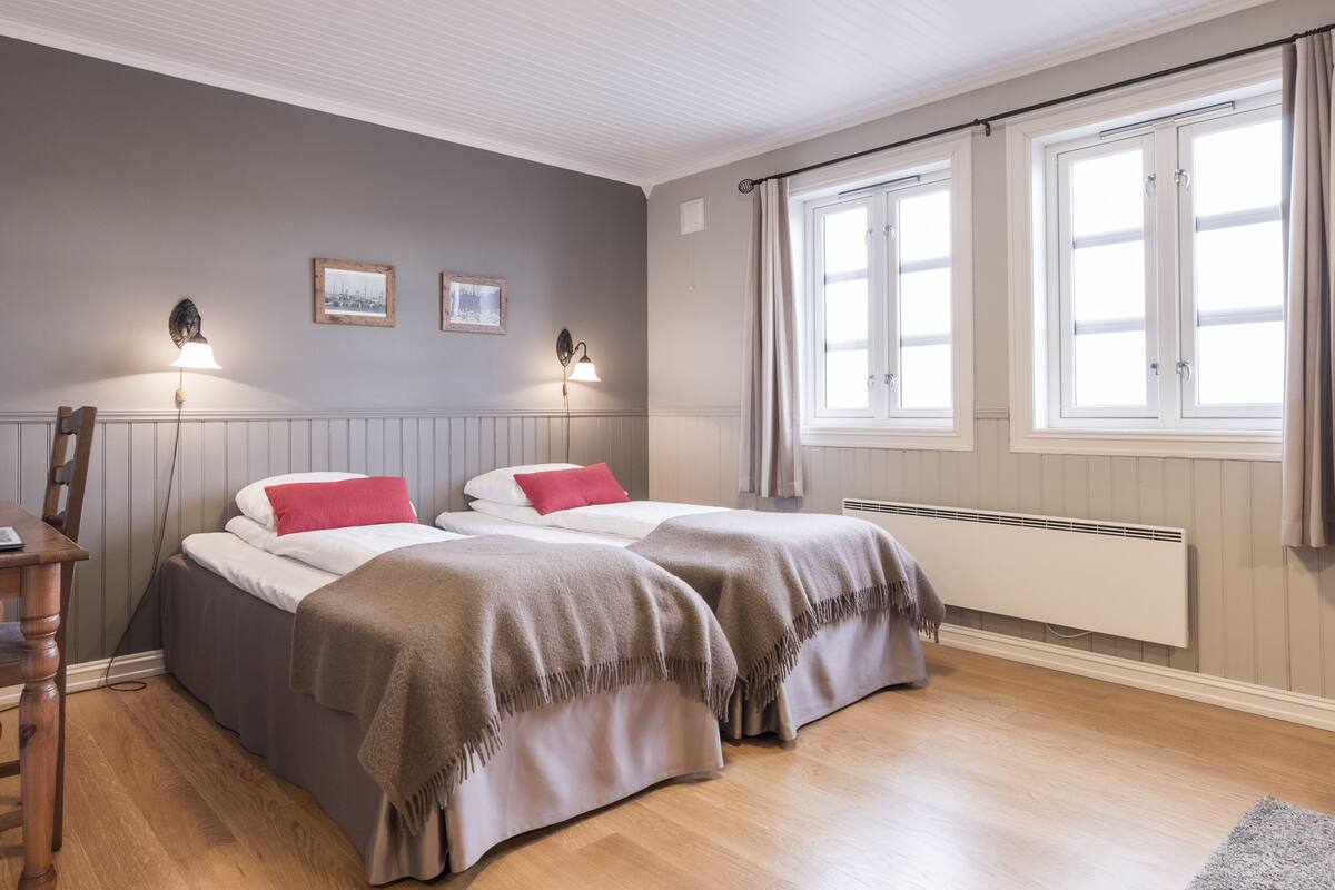 Habitación con dos camas, un escritorio y una silla. Cuenta con dos lámparas de pared y dos ventanas con cortinas.