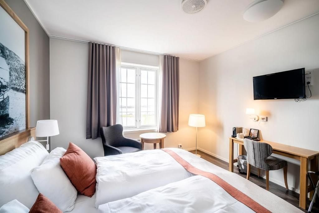 Quarto de hotel com cama, escrivaninha, cadeira, televisão e janela com cortinas.