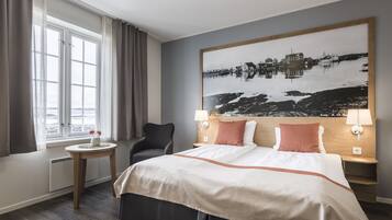 Habitación de hotel con una cama, una silla, una mesita con un florero, una ventana con cortinas y un cuadro colgado en la pared.