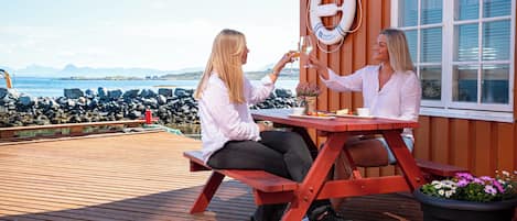 Two people sitting at a wooden picnic table with a lifebuoy on the wall, overlooking a rocky coastline.