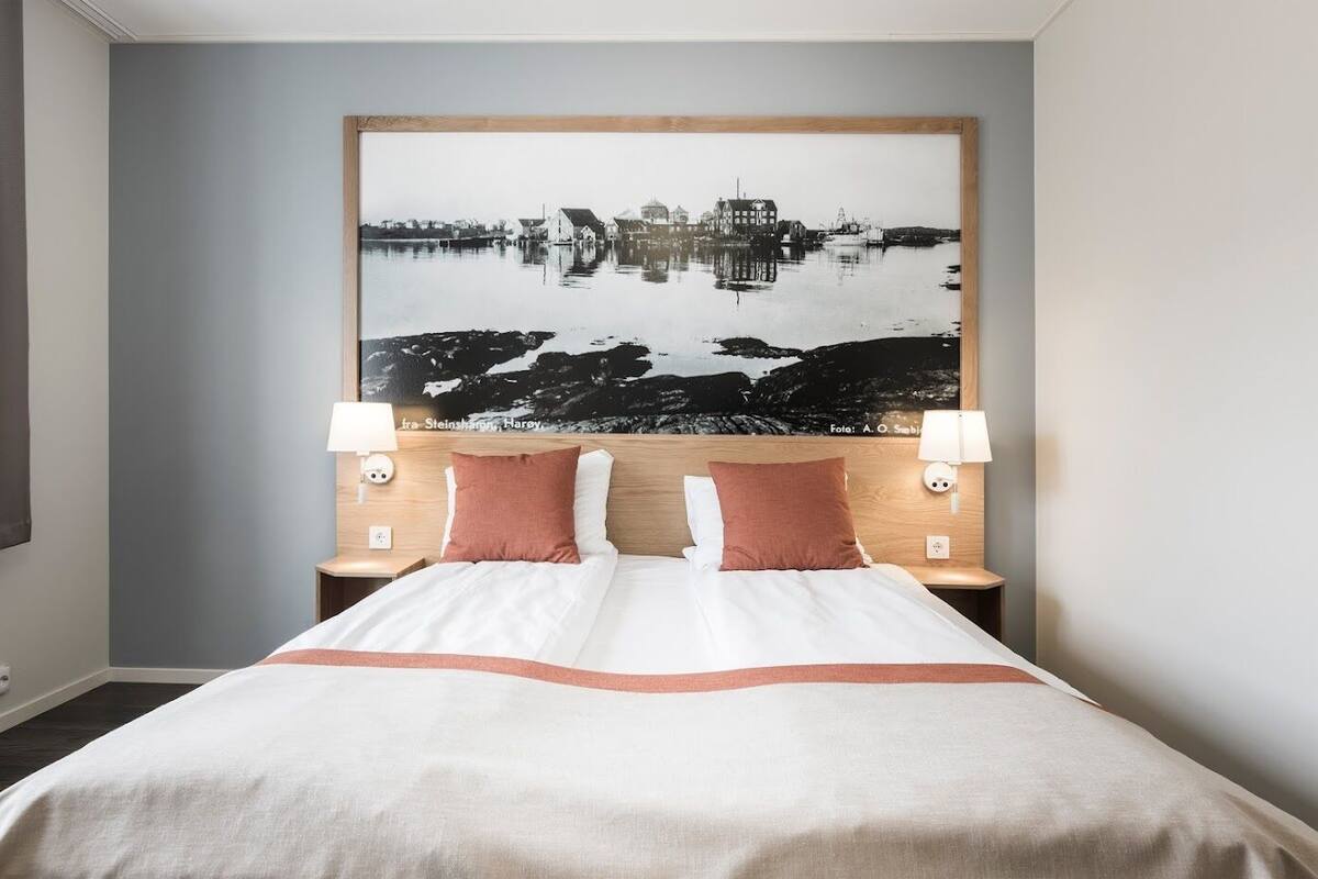 Habitación de hotel con una cama grande, dos lámparas de noche y una obra de arte en la pared que representa un paisaje.