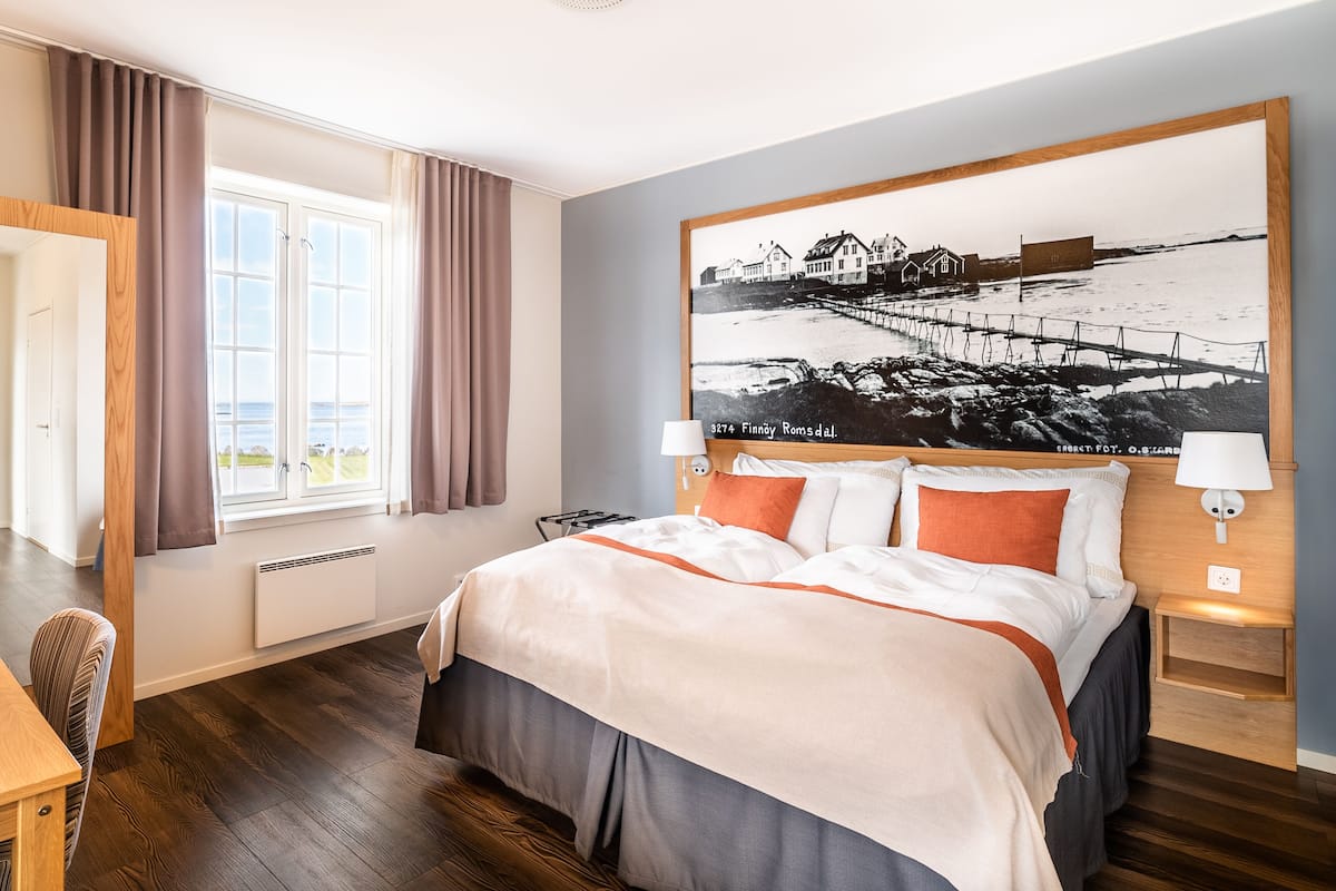 Habitación de hotel con una cama grande, una ventana con cortinas, un cabecero de madera y un cuadro colgado en la pared que muestra una escena costera.