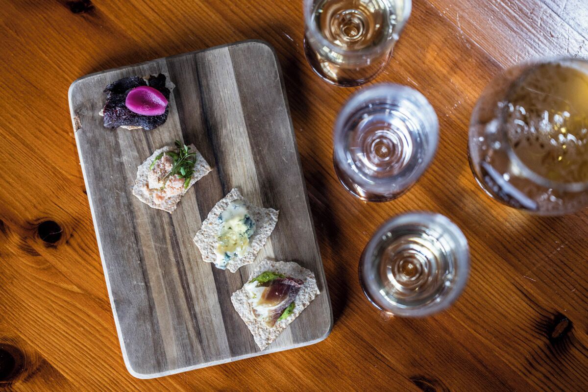 A wooden table with a rectangular serving board holding four pieces of food, accompanied by three glasses of a clear liquid.