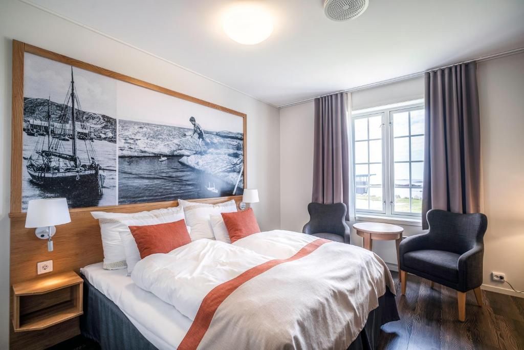 A hotel room with a bed, two chairs, a small table, a window with curtains, and artwork on the wall.