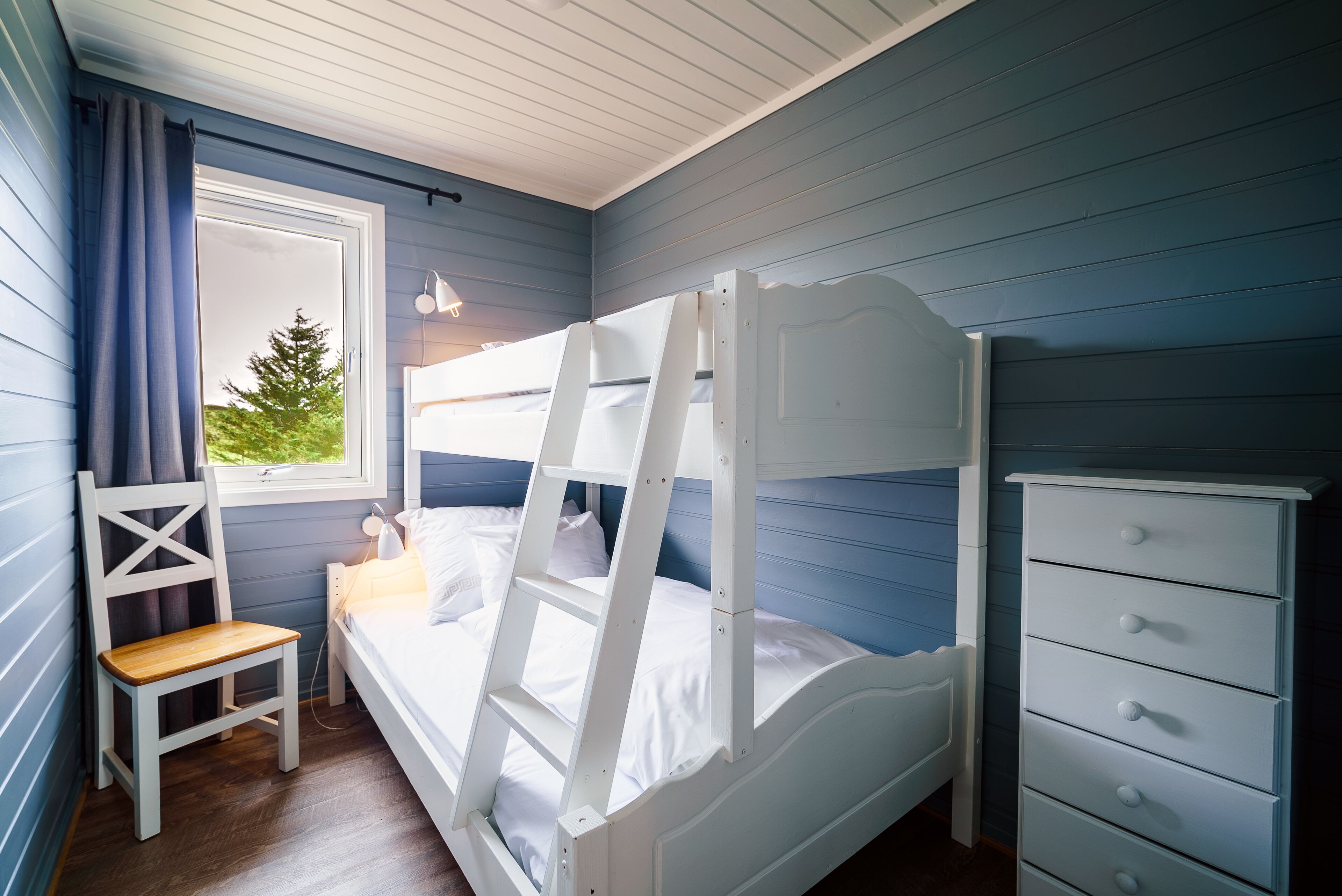 A bunk bed with a ladder, a wooden chair, a dresser, and a window with curtains.