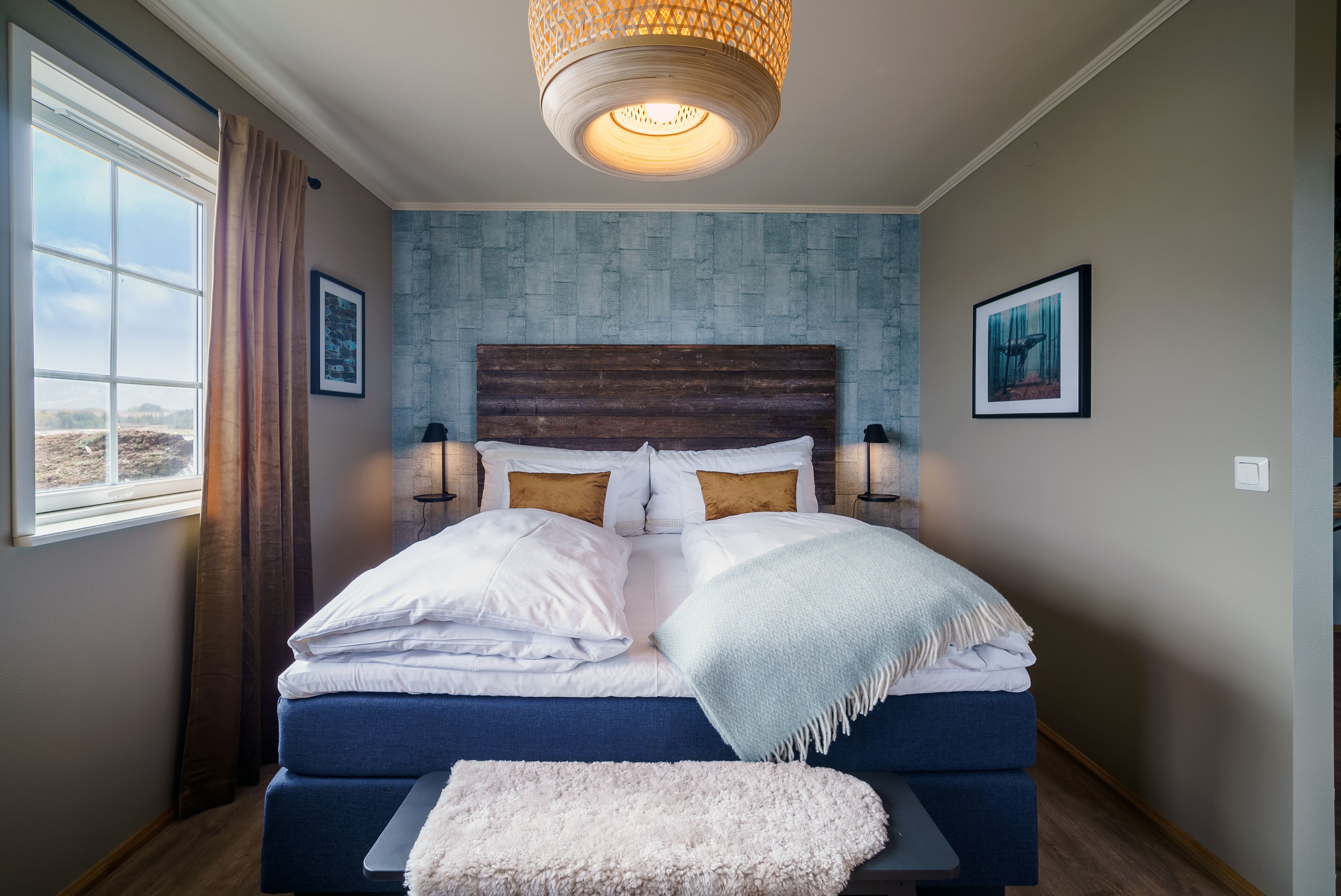 A bedroom with a large bed, a wooden headboard, two bedside lamps, a window with curtains, and a view of the outdoors.