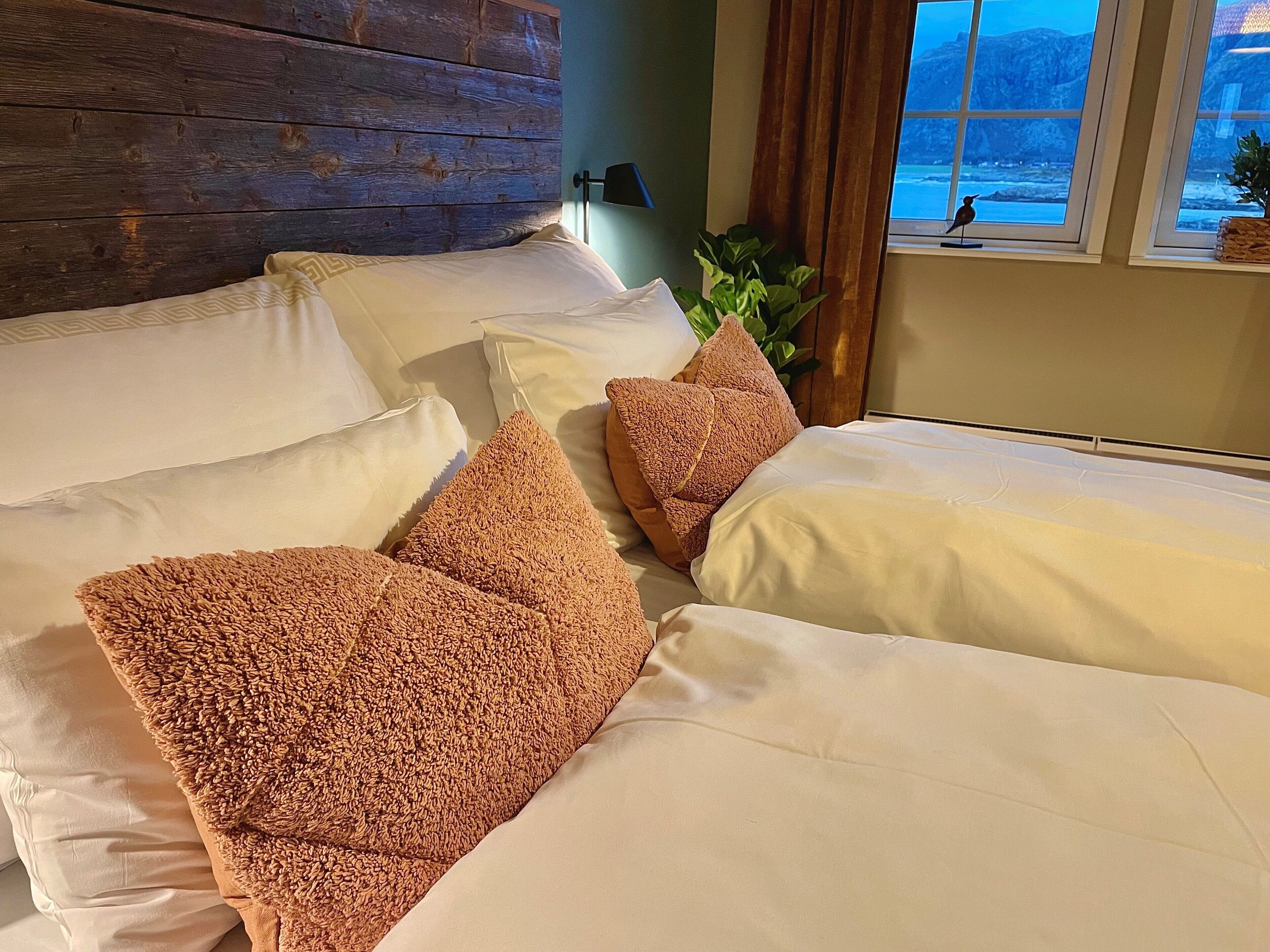 Una cama con ropa blanca y almohadas marrones con textura, un cabecero de madera, una ventana con vistas a montañas y una planta en el alféizar.
