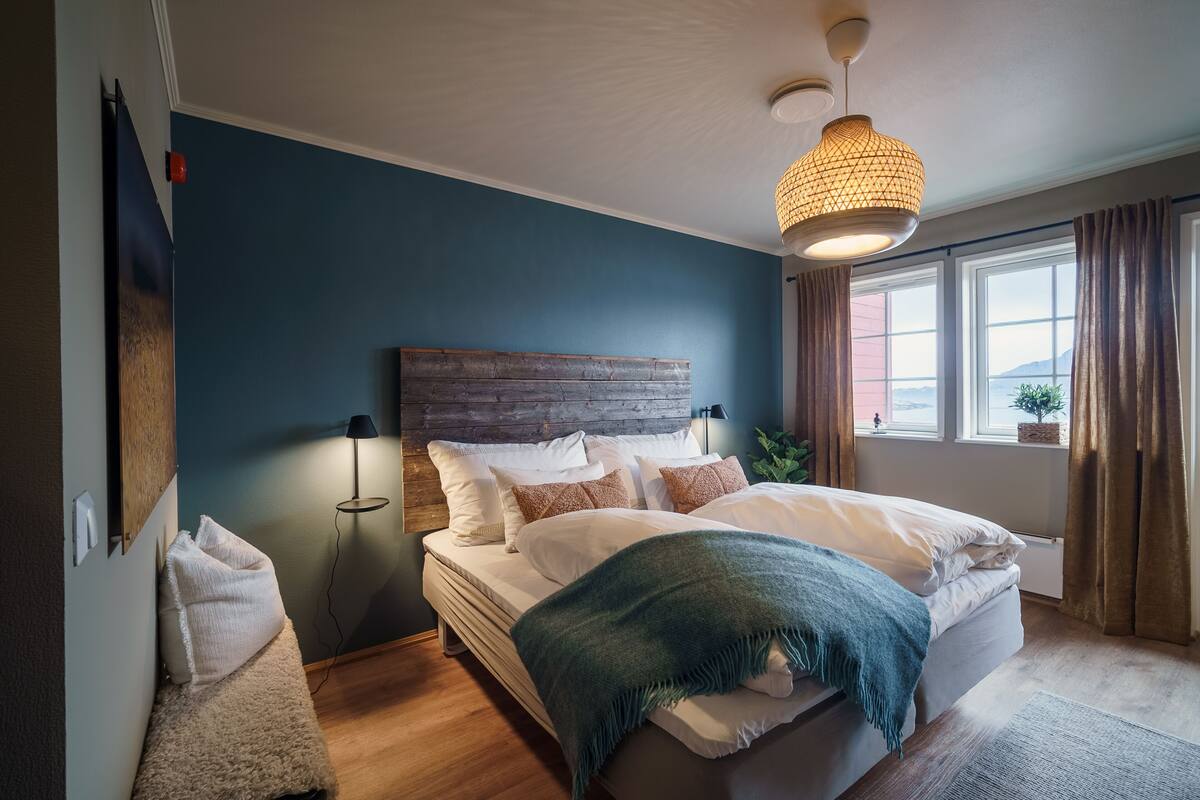 A bedroom with a large bed, a wooden headboard, a hanging light fixture, and a view of the outdoors through a window.