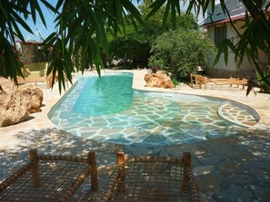 A swimming pool with a stone pattern, surrounded by rocks and plants, with a wooden fence in the foreground. - Kamili View Apartment in Zanzibar (Kiwengwa)