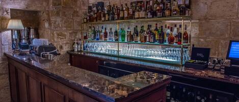 Una zona de bar con un fondo de pared de piedra, una barra de madera y estantes llenos de diferentes botellas de licor.
