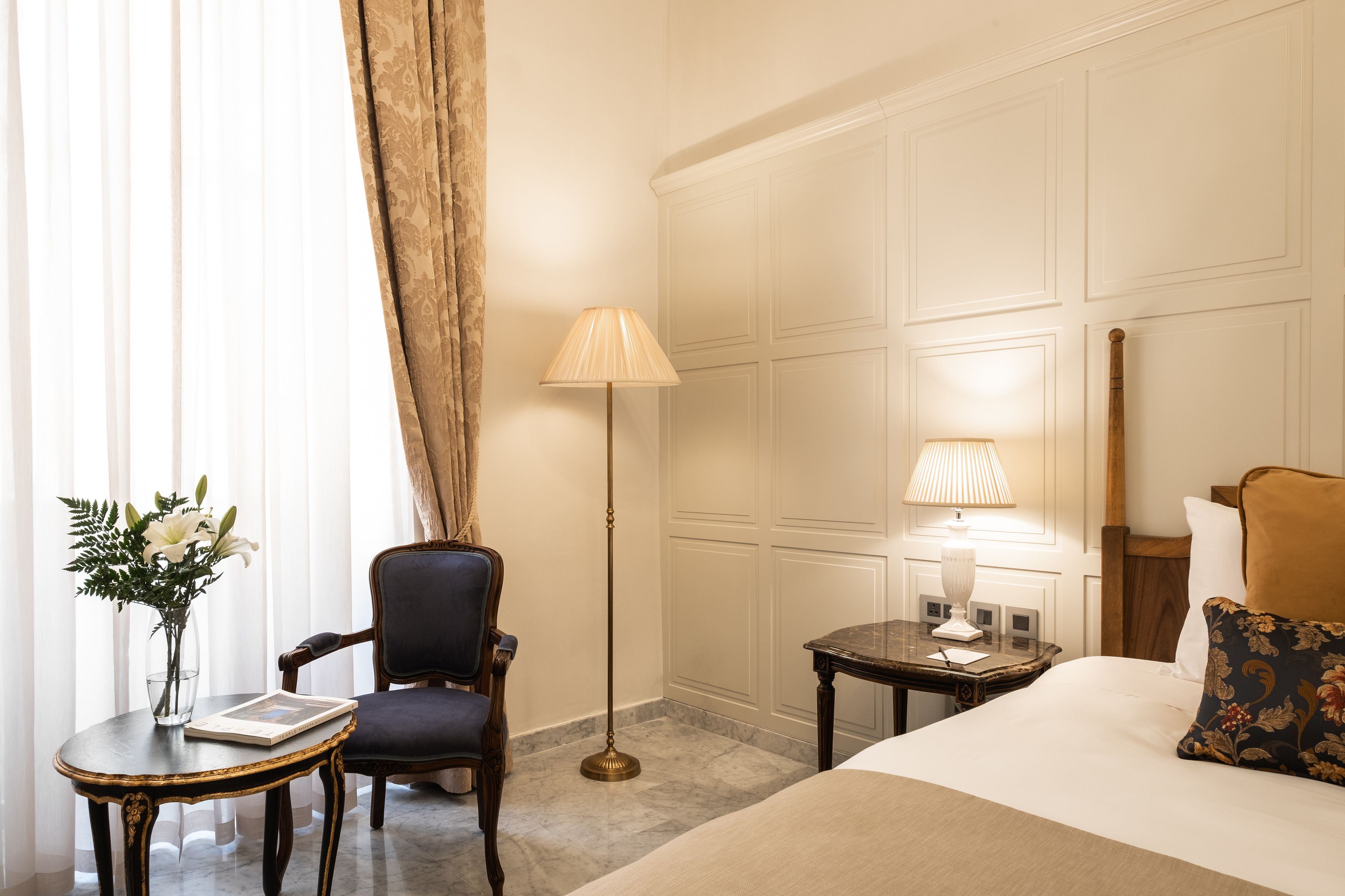 Ein Hotelzimmer mit einem Bett, einem Sessel, einem kleinen Tisch mit Blumenvase, einer Stehlampe und einem Nachttisch mit Lampe.