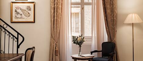 A hotel room with a bed, a desk with drawers, a chair, a small table with a vase of flowers, and a lamp.