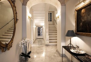 Un majestuoso pasillo con suelos de mármol, un espejo grande y ornamentado, un jarrón con orquídeas blancas y un cuadro en la pared.