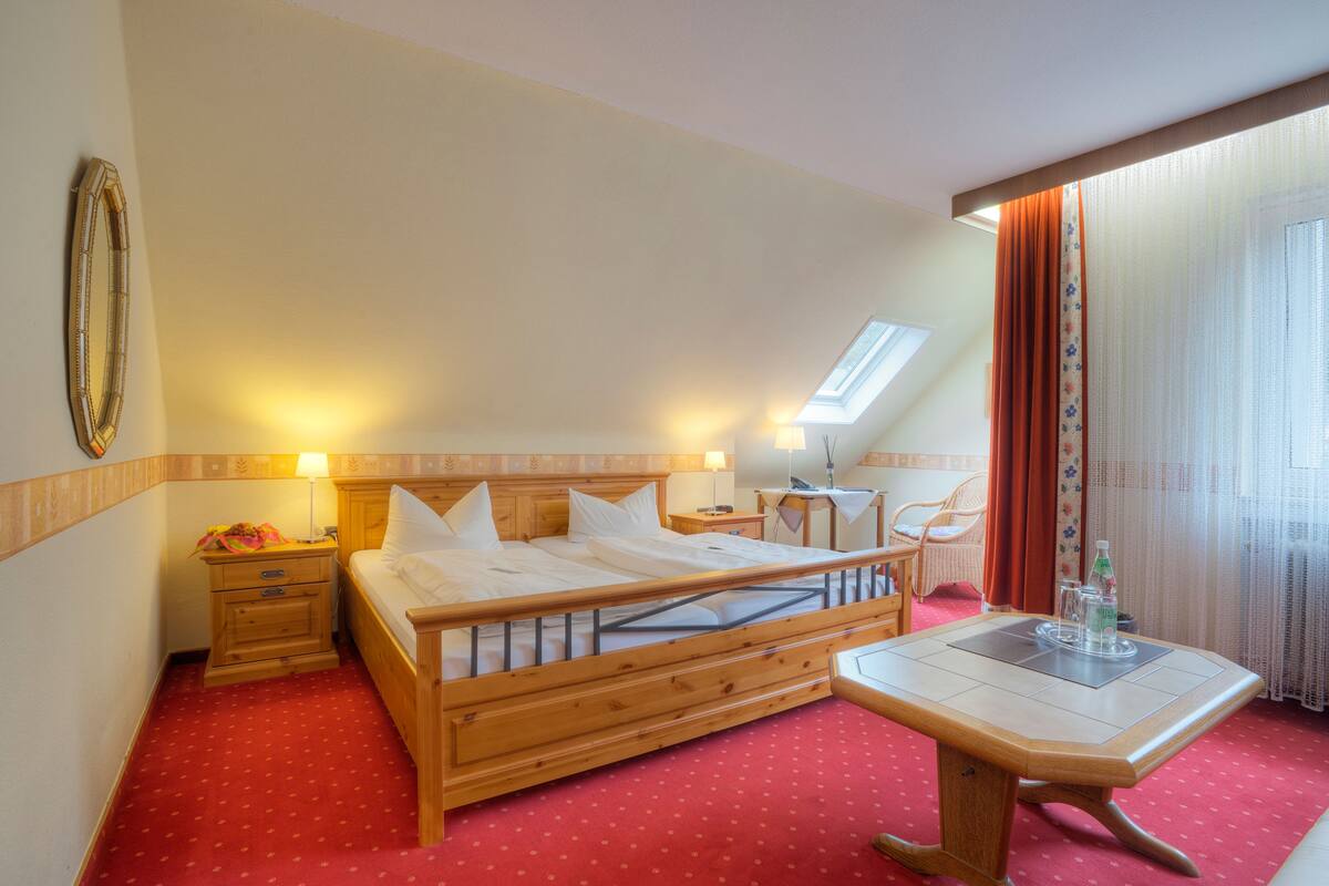 Habitación de hotel con cama de madera, una mesita, una mesita de noche, una ventana con cortinas y una lámpara de pared.