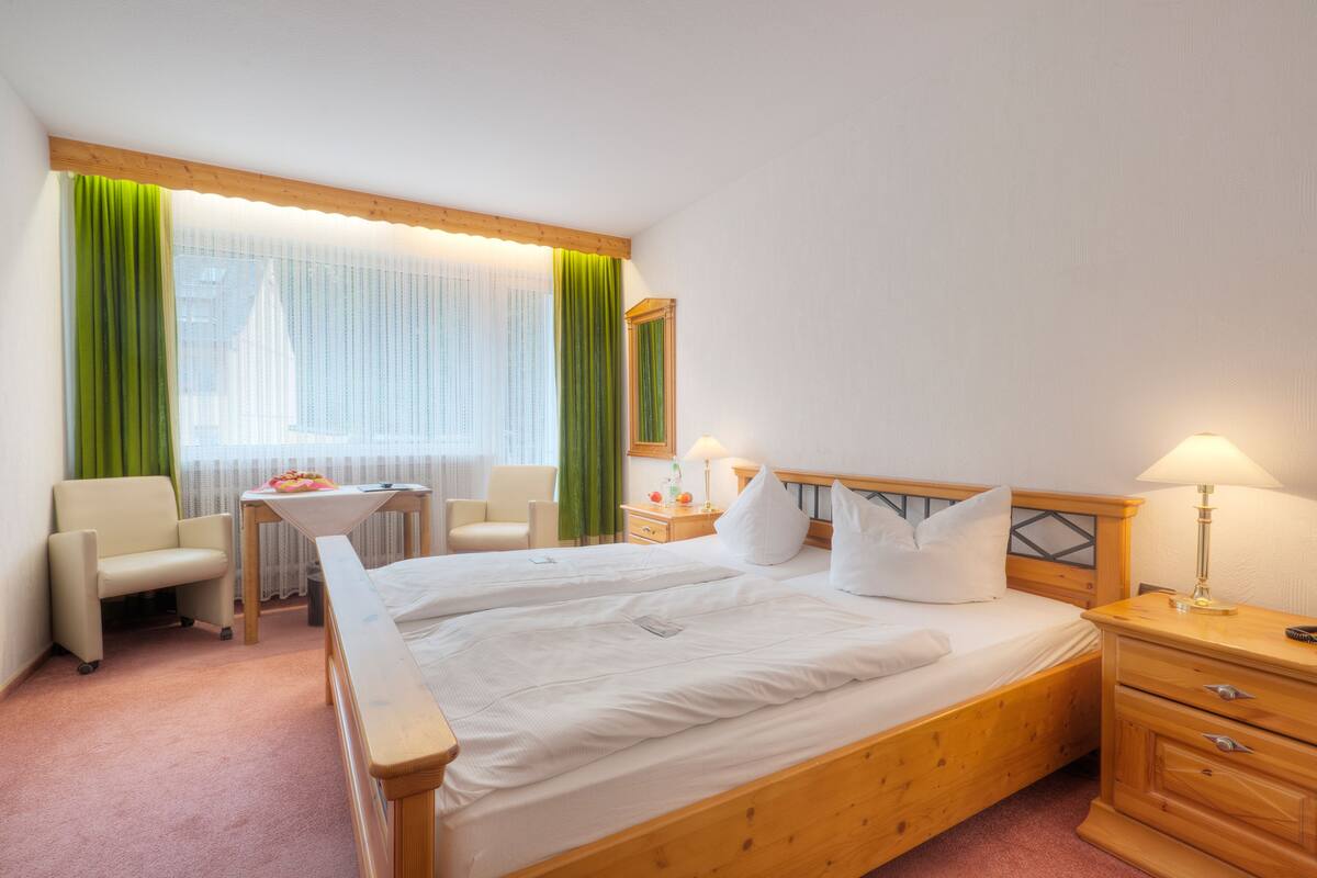 Habitación de hotel con una cama grande, dos mesitas de noche, una silla, una pequeña mesa y una ventana con cortinas.