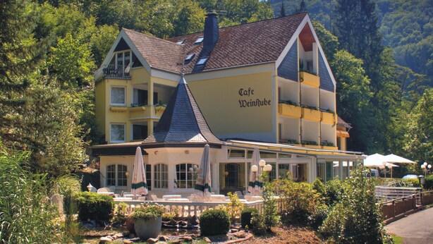 Een meerhoog gebouw met een café genaamd "Cafe Weinmube" op de begane grond, omringd door weelderig groen en een kleine tuin.