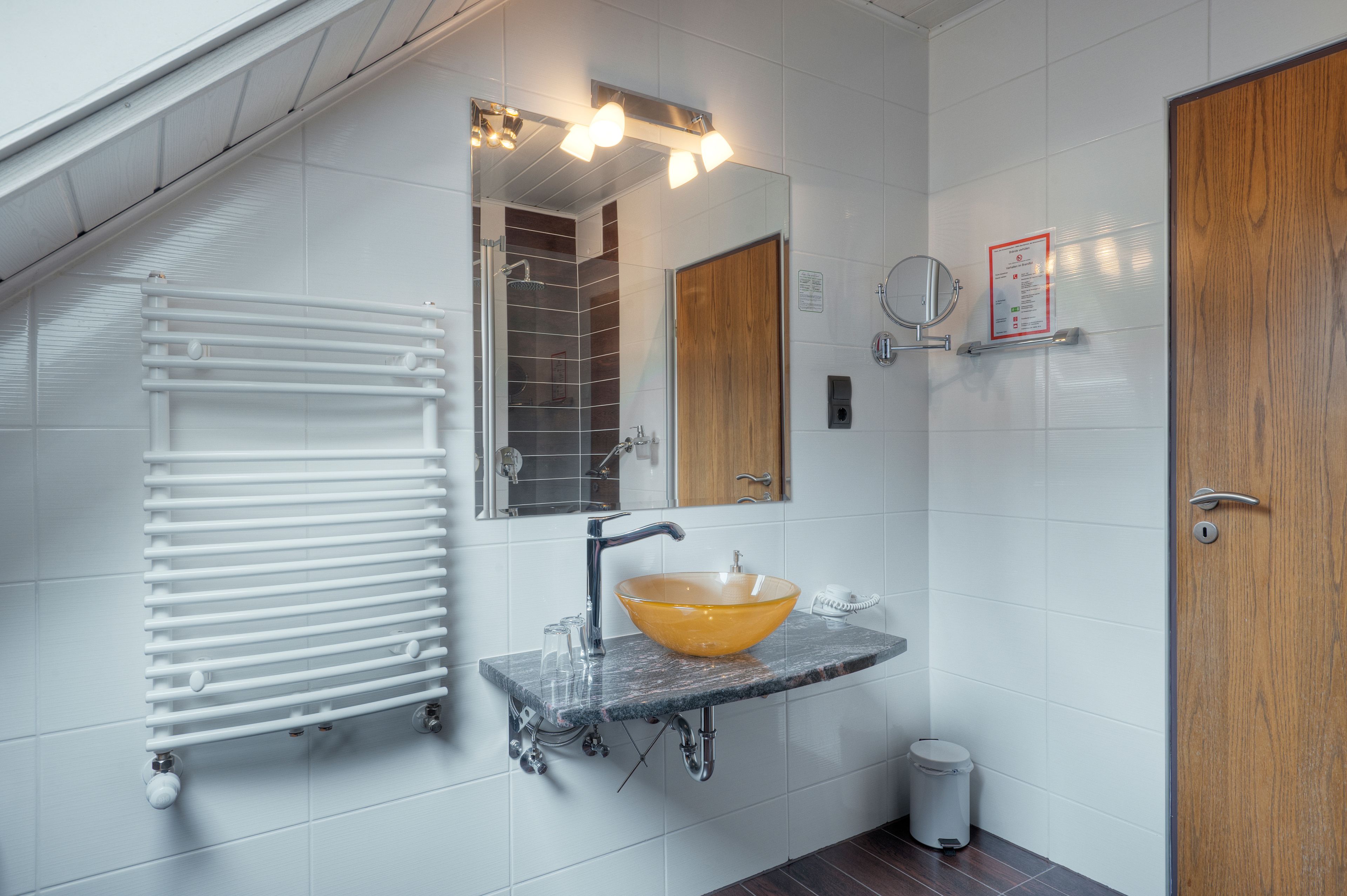 Een badkamer met een witte betegelde muur, een houten deur, een glazen wastafel met een gele kom, een spiegel en een witte radiator.