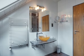 Un baño con pared de azulejos blancos, puerta de madera, lavabo de cristal con un lavamanos amarillo, espejo y radiador blanco.
