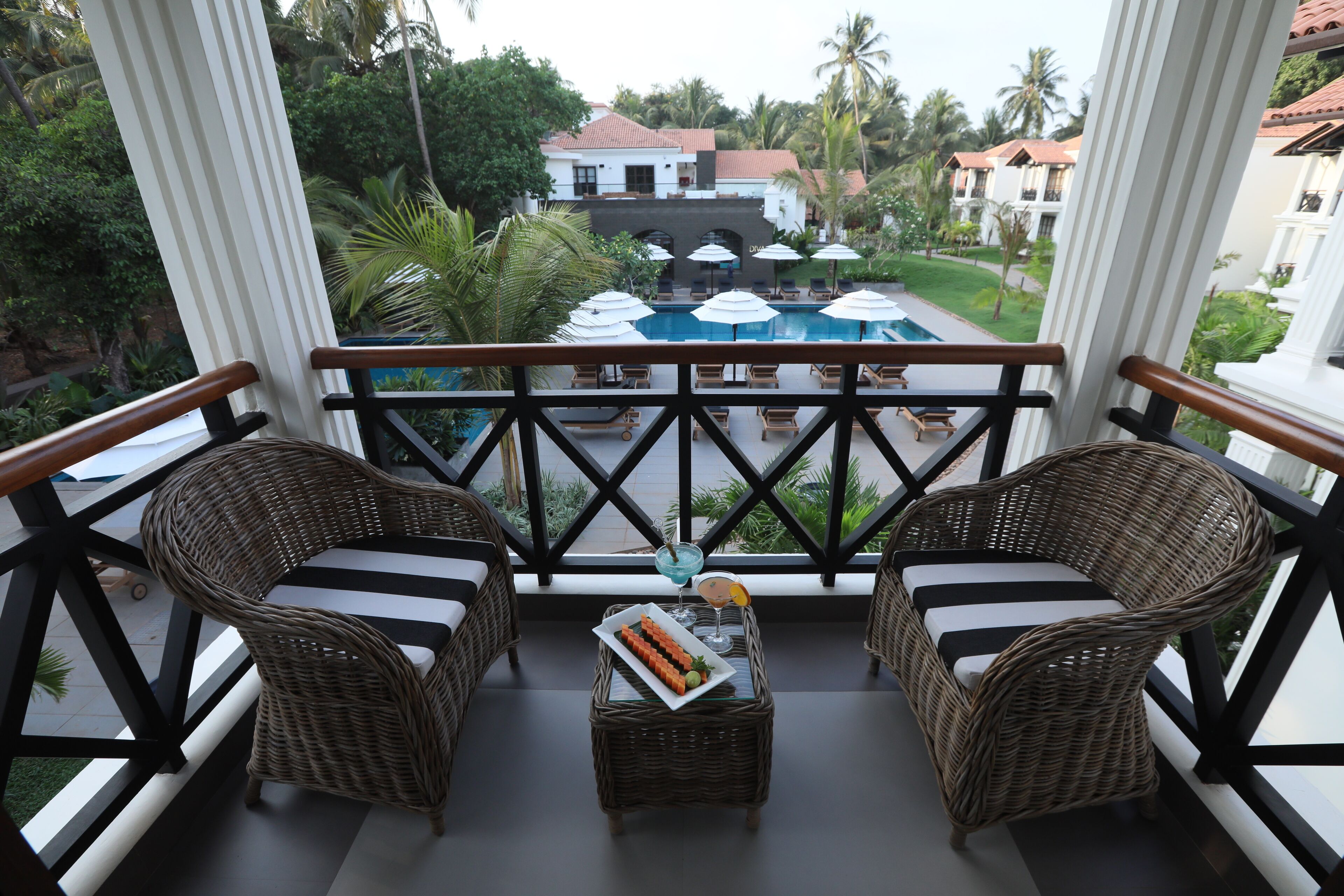 A balcony with wicker furniture, a small table with a tray of food, and a view of a pool and buildings.
