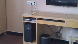 Una habitación con una televisión montada en la pared, un escritorio de madera con una computadora y un cesto de basura debajo, y un enchufe en la pared.