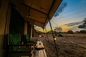 Un área de comedor al aire libre con carpas, mesas y sillas, vista a la sabana al atardecer y un cielo despejado.