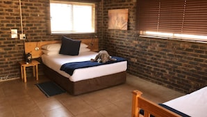 A bedroom with a brick wall, a bed with a blue blanket, a wooden nightstand, and a window with blinds. - Zingela Nature Reserve - Doornstock Lodge (Blouberg)