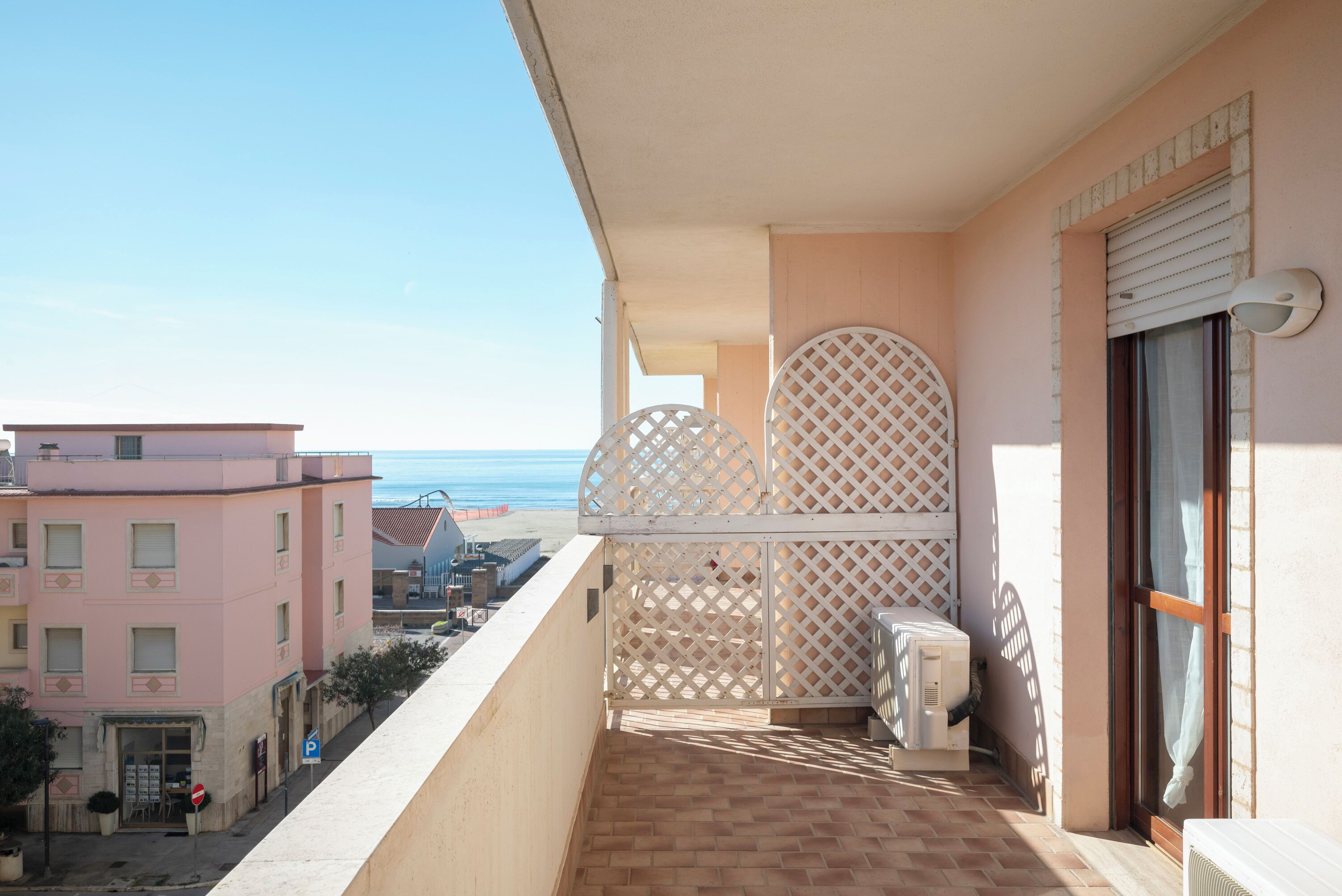 Un balcone con vista sulla spiaggia, un parapetto in legno chiaro e un edificio dalla facciata rosa.
