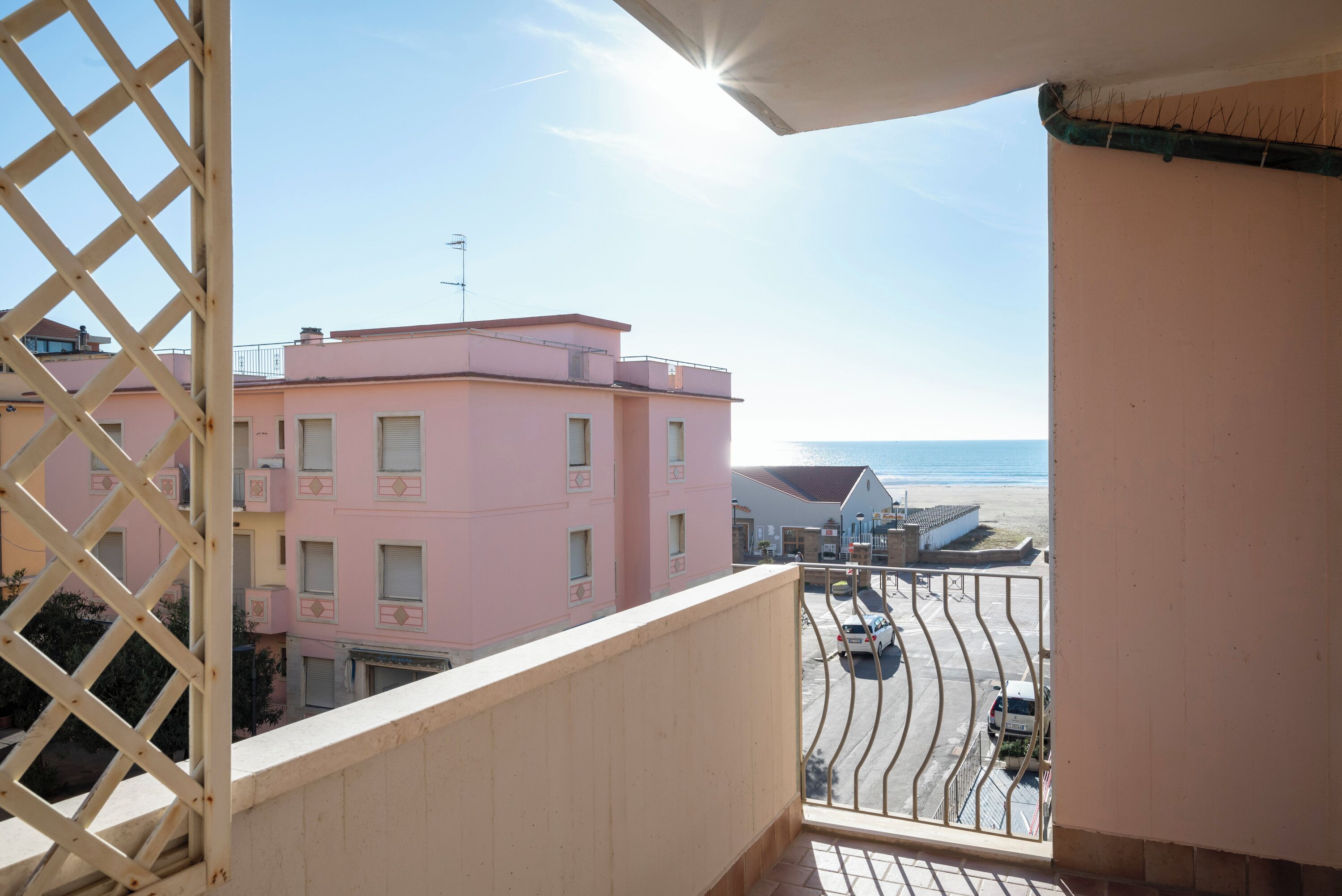 Un balcone con vista su un edificio rosa, una strada con auto parcheggiate e un cielo sereno.