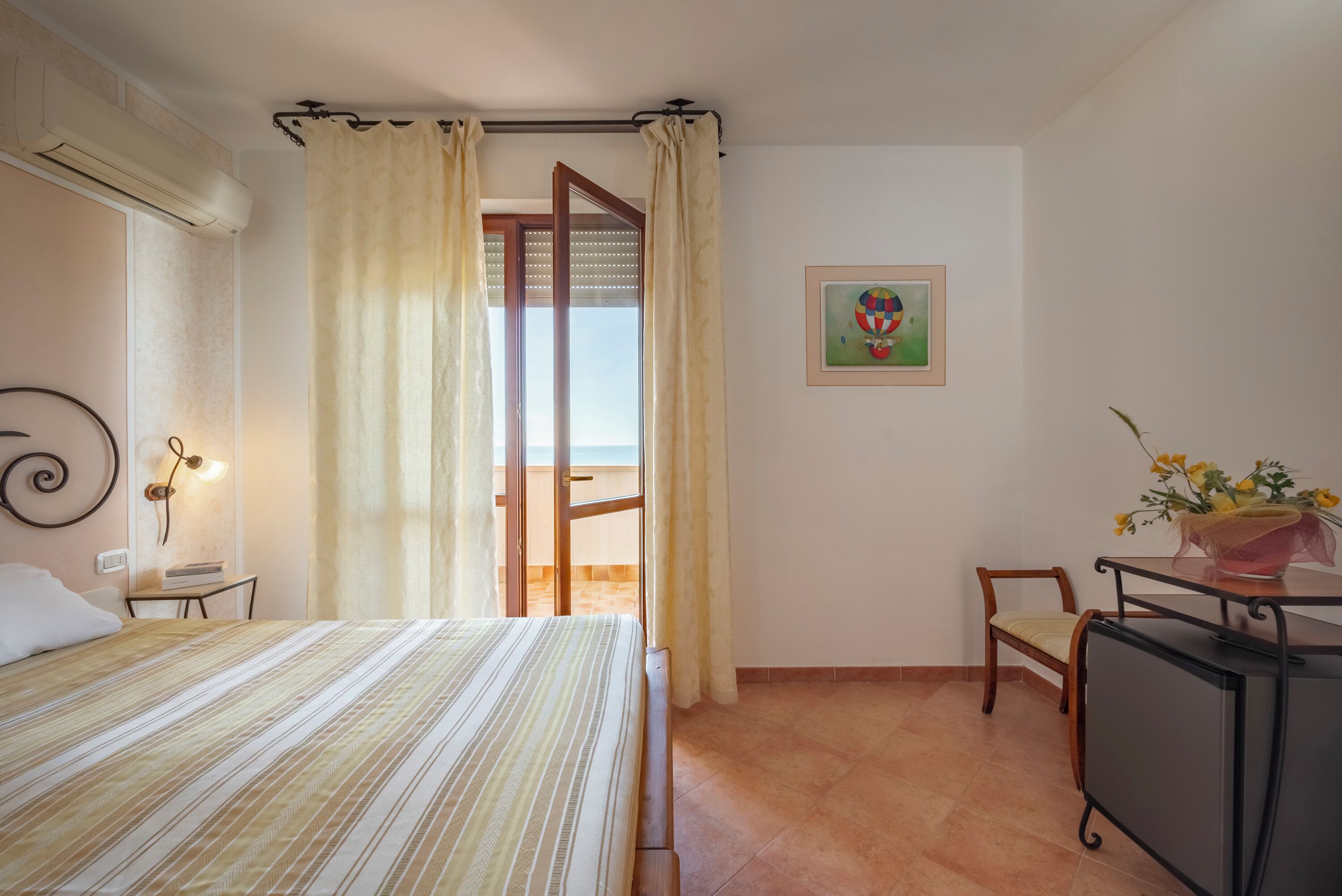 Un dormitorio con cama, una silla, un escritorio, aire acondicionado instalado en la pared, una puerta con cortinas, y un cuadro enmarcado en la pared.