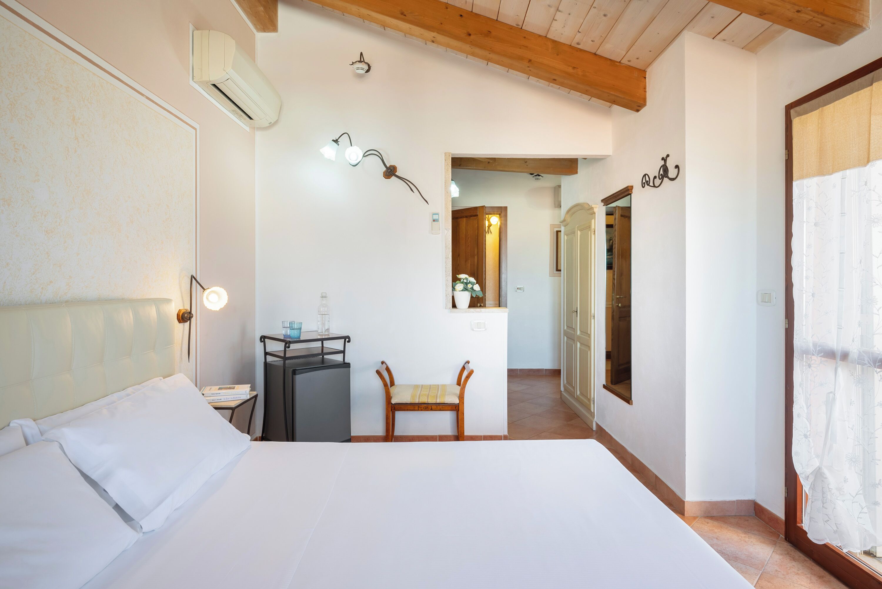 Ein Schlafzimmer mit einem Bett, Nachttisch und einem Sessel. An der Wand befindet sich ein Spiegel und eine Tür zu einem anderen Raum.