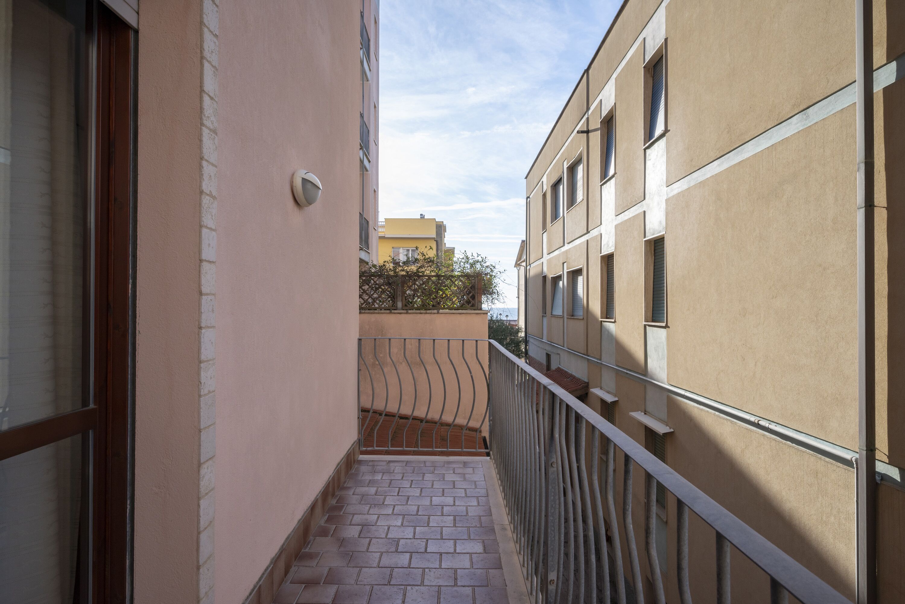 Un balcón estrecho con barandilla de metal, suelo de ladrillo y vista a edificios cercanos.
