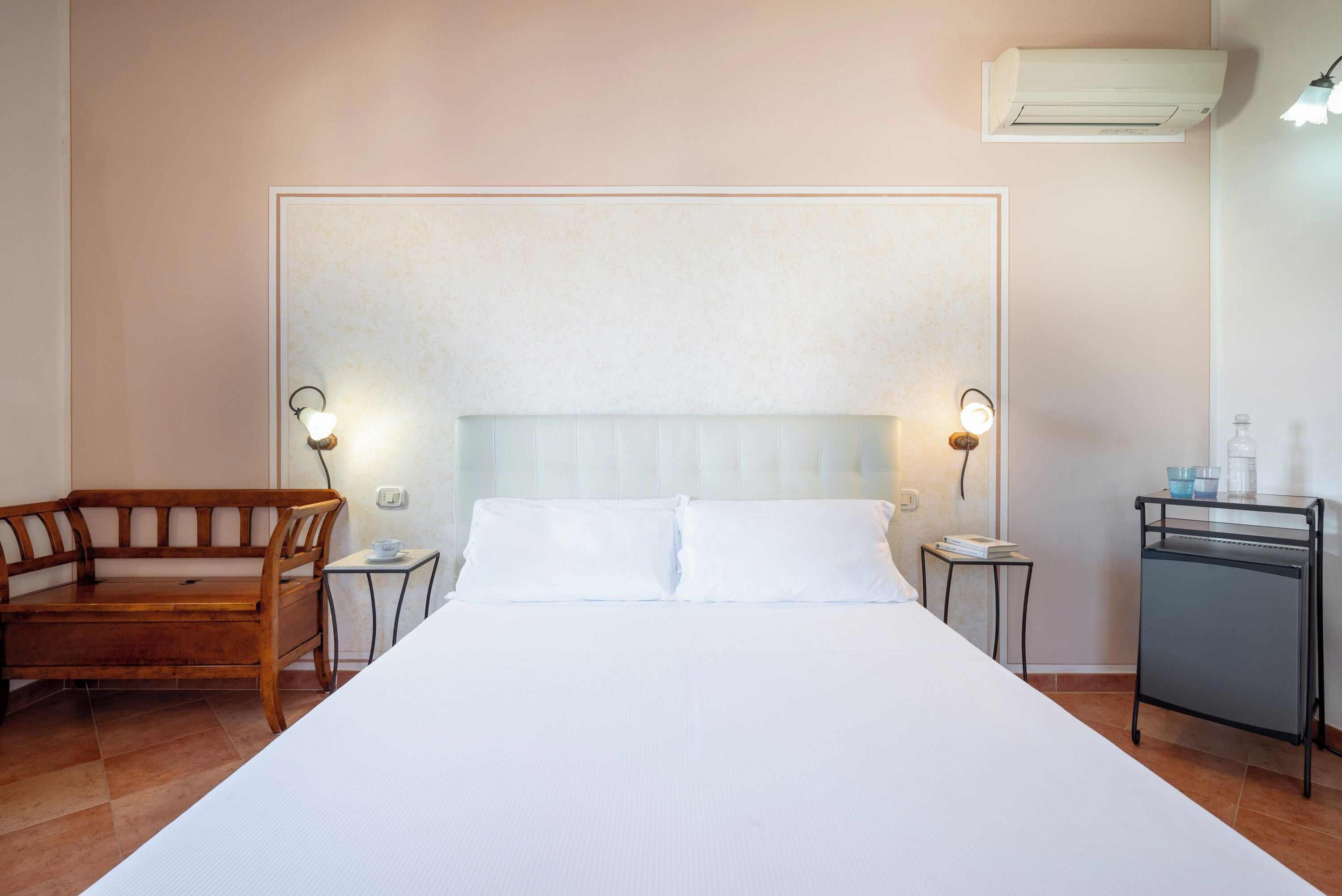 A hotel room with a large bed, a wooden bench, a small table, and a nightstand with a lamp.